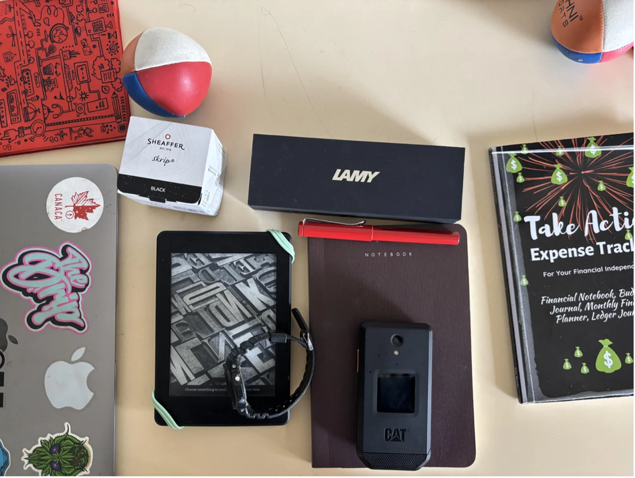 Everyday carry layout shared to r/dumbphones — juggling ball, Sheaffer ink, Lamy pen box, e-reader, CAT flip phone.